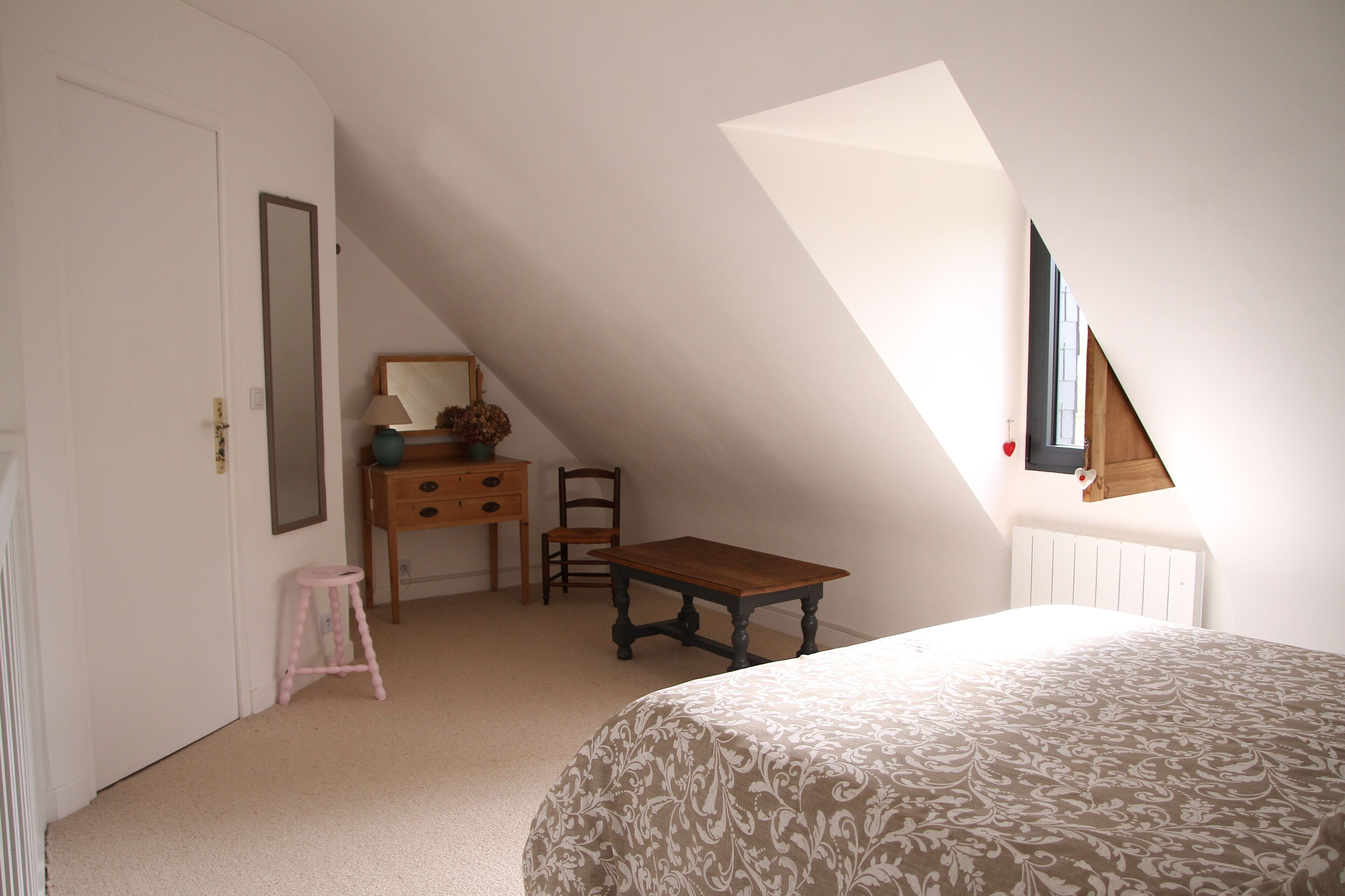 A bedroom with a sloped ceiling, a bed, a dresser, a chair, and a small stool.