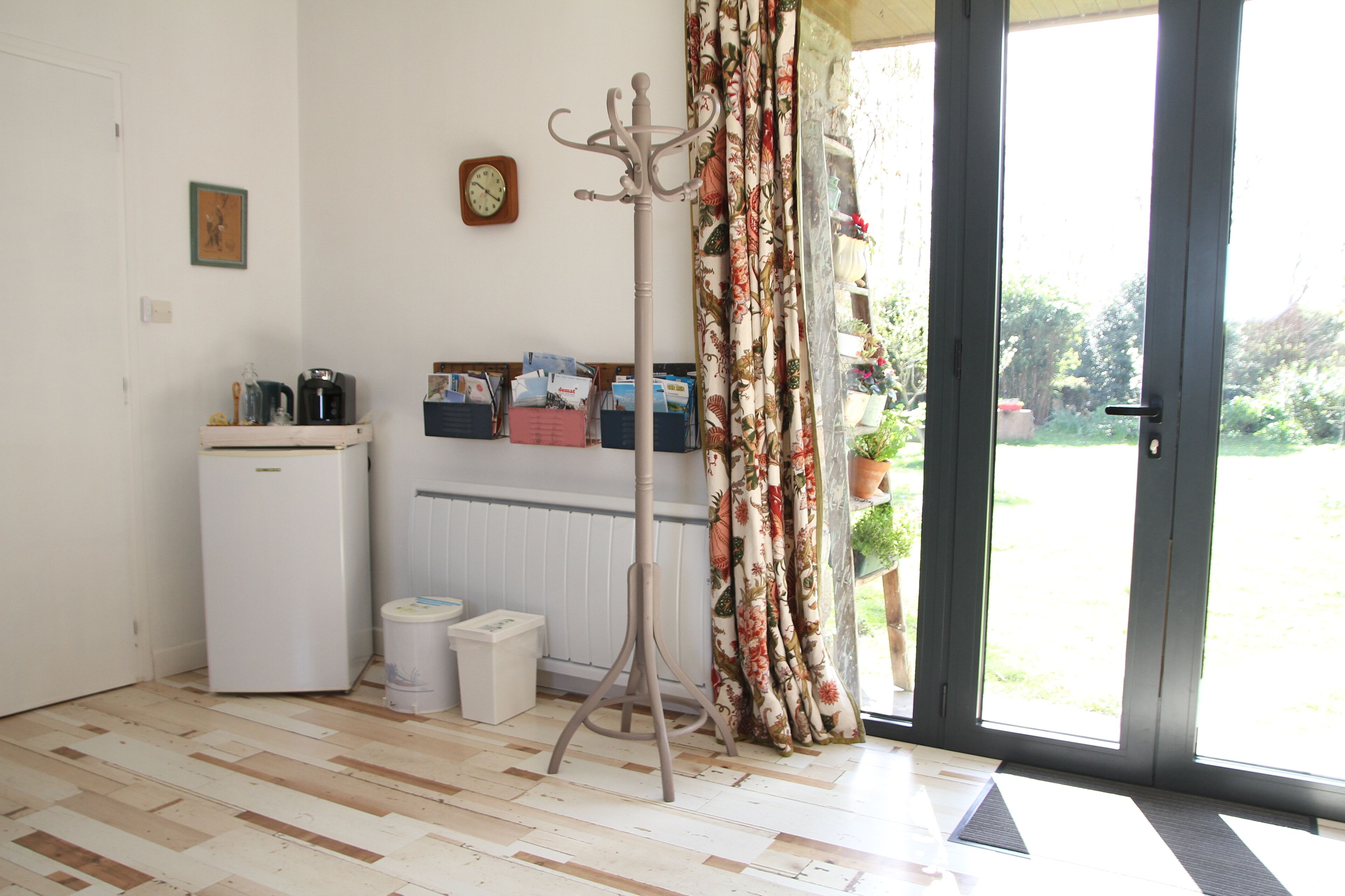 A room with a wooden floor, a white radiator, a small white fridge, a coat stand, a door with a view to the outside, and a wall with a clock and picture.