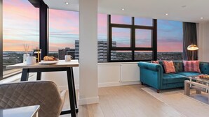 Un salon moderne doté d’une grande fenêtre donnant sur la ville, d’un canapé bleu, d’une table à manger avec un croissant et d’une lampe.