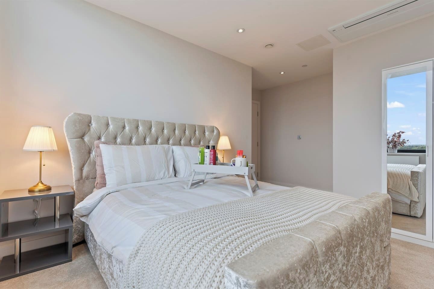 A modern bedroom with a tufted headboard, a bedside table with a lamp, a bedside table with toiletries, and a view through a glass door.