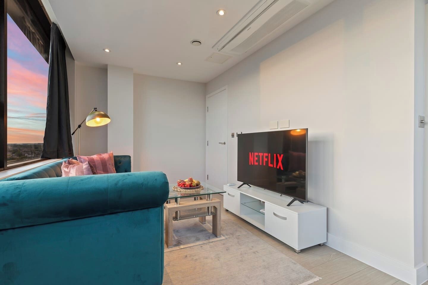 A modern living room with a large flat-screen TV displaying the Netflix logo, a teal sofa, a small coffee table with fruit, and a view of the city through a large window.