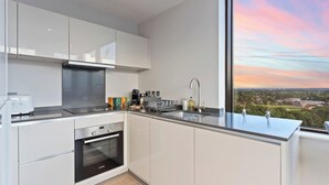 Une cuisine moderne avec des armoires blanches, un four encastré, une hotte de cuisinière en acier inoxydable et une grande fenêtre offrant une vue sur le paysage urbain au coucher du soleil.