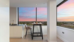 Une cuisine moderne dotée d’une grande fenêtre, un coin repas avec une table et des chaises, et une vue sur le coucher de soleil au-dessus d’un paysage urbain.