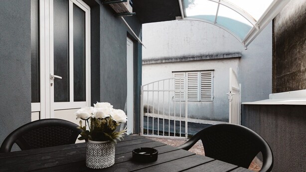 Une terrasse avec une table, des chaises, un vase de fleurs et une vue sur un bâtiment avec des volets.