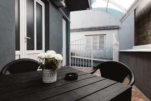 Une terrasse avec une table, des chaises, un vase de fleurs et une vue sur un bâtiment avec des volets.