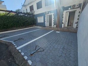 Une cour dallée, agrémentée d’un portail en métal, d’une haie et d’un bâtiment doté d’une porte et de fenêtres.