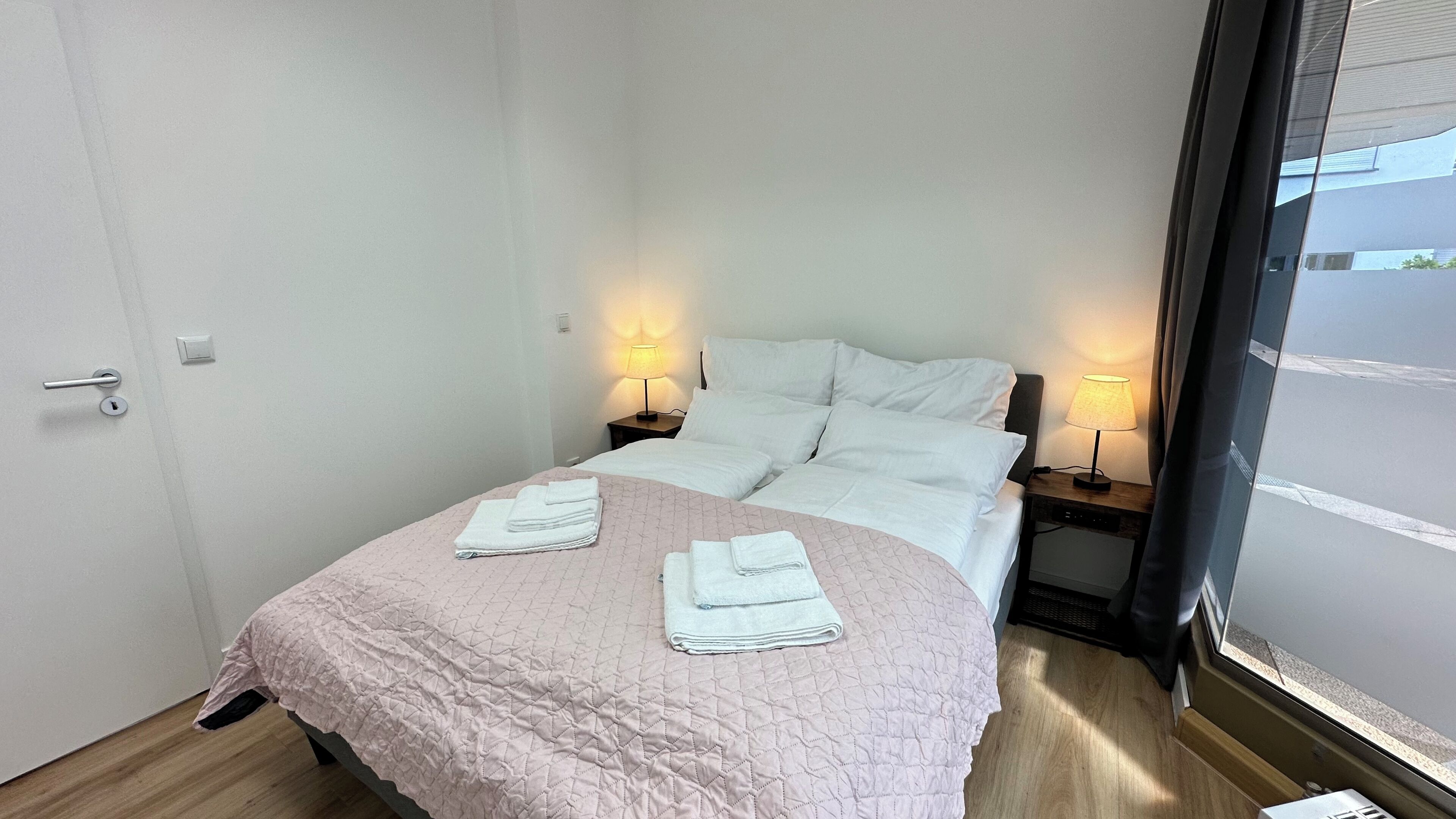 A hotel room with a bed, two bedside tables with lamps, a door, and a window.