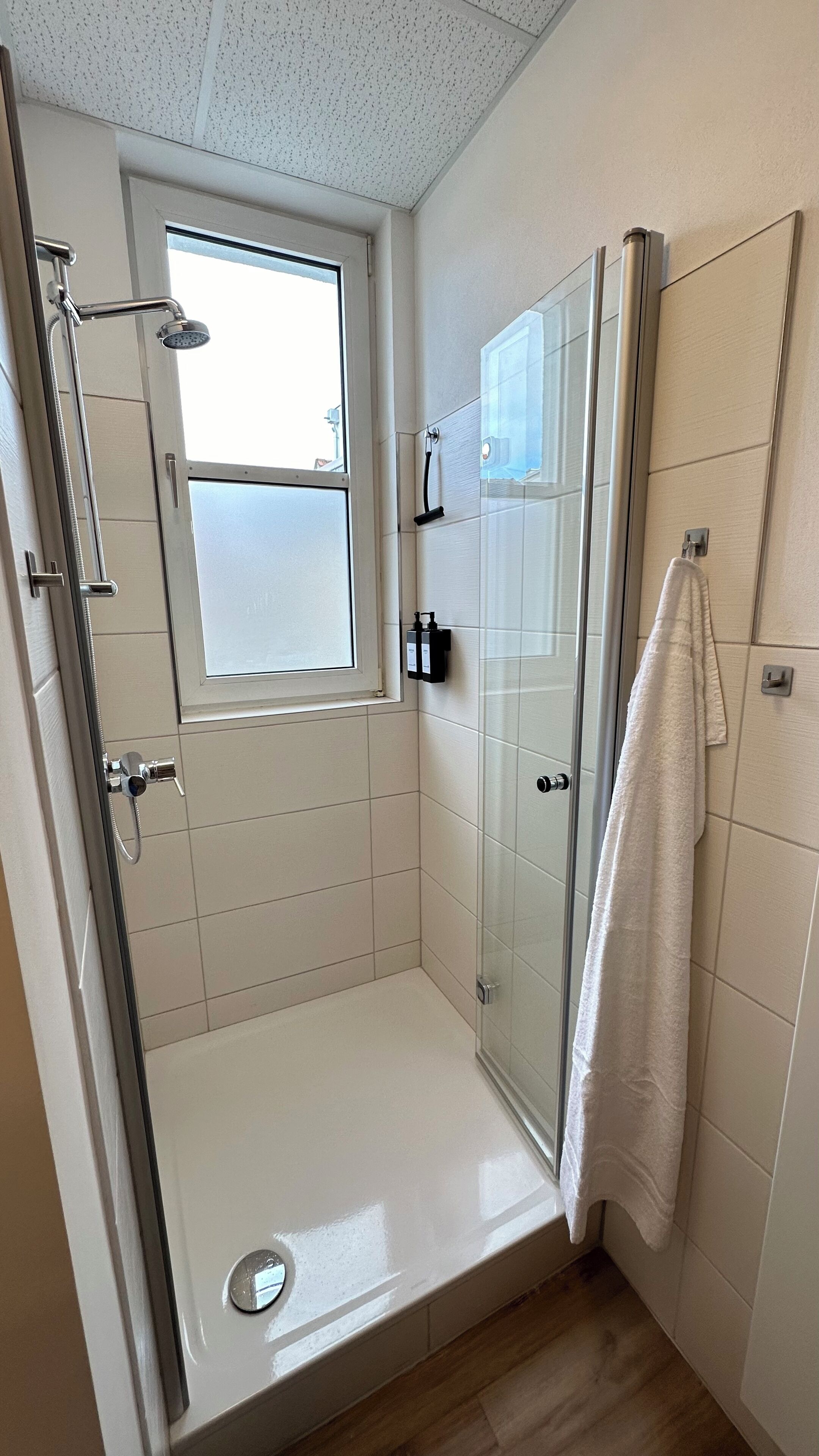 Een kleine, moderne douchekamer met een glazen deur, een raam en een handdoek aan de muur.