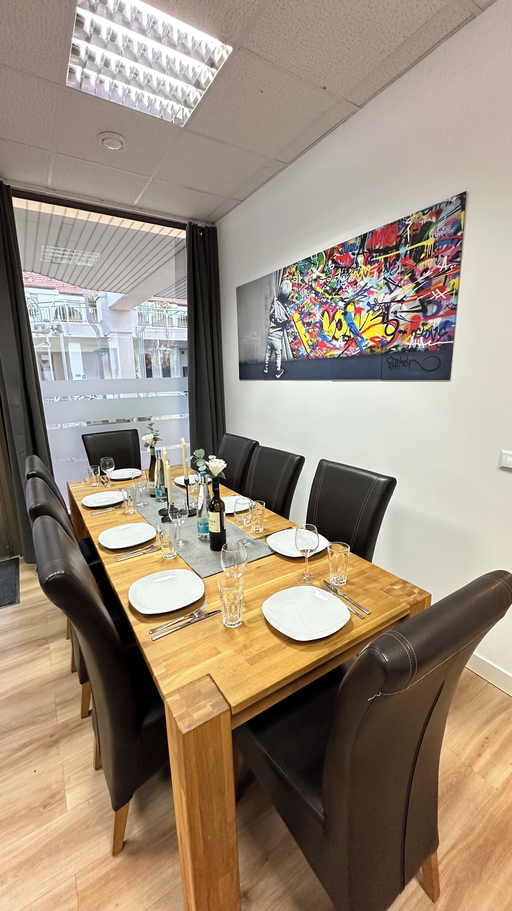 Un coin repas est aménagé avec une longue table en bois, dressée pour six personnes, et entourée de chaises en cuir noir. Un tableau abstrait aux couleurs vives orne le mur au-dessus de la table.