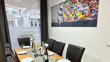 Un coin repas est aménagé avec une longue table en bois, dressée pour six personnes, et entourée de chaises en cuir noir. Un tableau abstrait aux couleurs vives orne le mur au-dessus de la table.