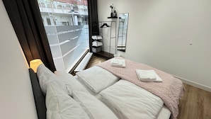 Un dormitorio con cama, ventana con vista a un edificio, radiador y un estante con objetos.