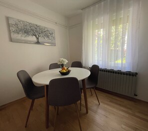 Un comedor con una mesa redonda, cuatro sillas, un cuenco de fruta y un florero. Hay un cuadro en la pared y una ventana con cortinas.