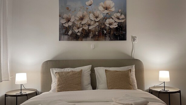 Ein ordentlich gemachtes Bett mit gefalteten Handtüchern, zwei Nachttischlampen und einem Blumenbild an der Wand.