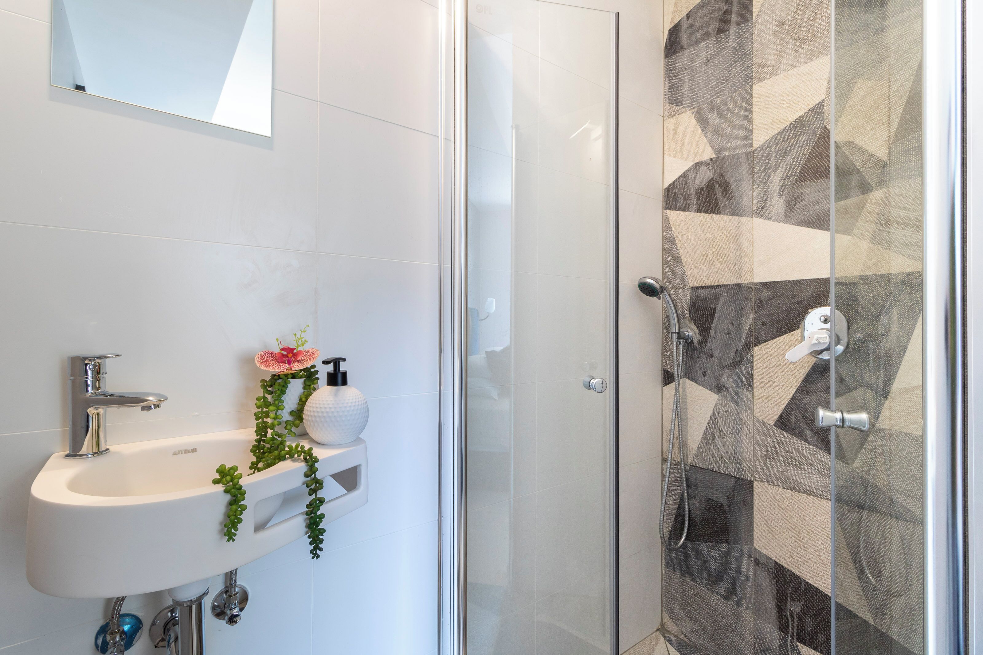 Un baño moderno con un lavamanos blanco, mampara de ducha de vidrio y pared de ducha con diseño geométrico.