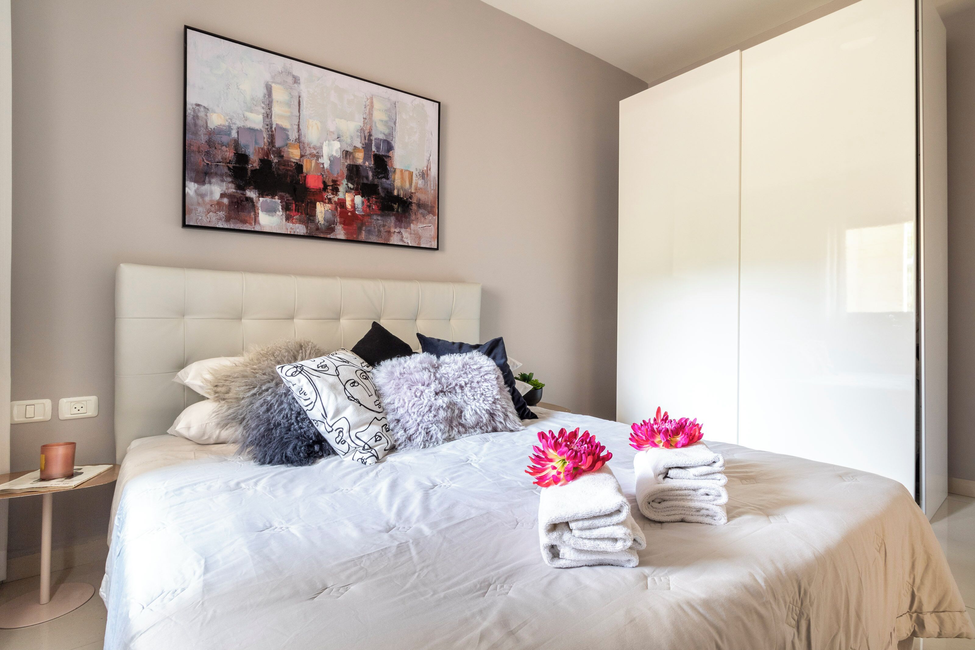 A modern hotel room with a large bed, a cityscape painting, and a wardrobe.