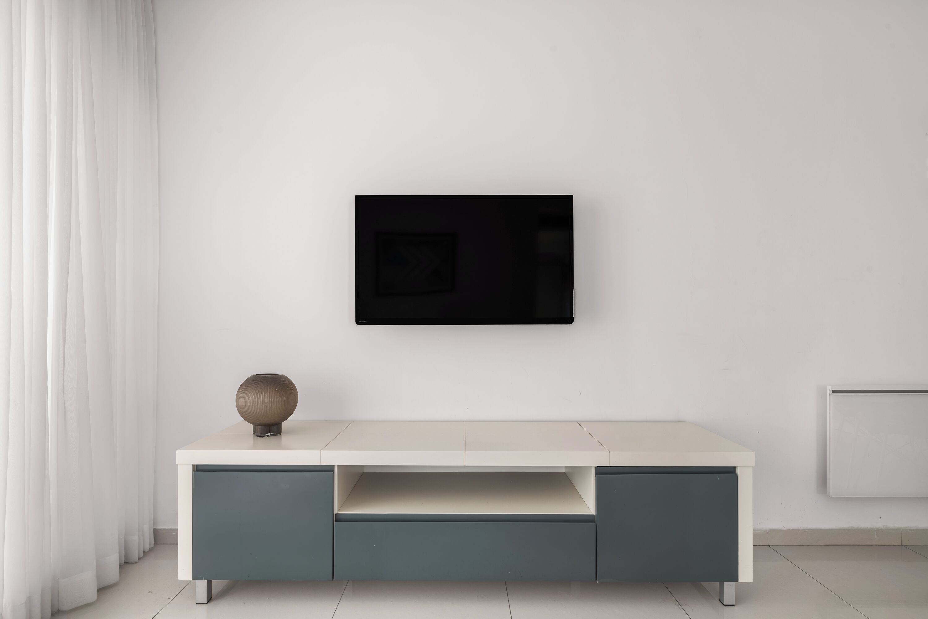 Een moderne, minimalistische kamer met een witte muur, een flatscreen televisie boven een lichtgekleurd console tafeltje met een decoratieve bol, en een witte radiator aan de rechterkant.