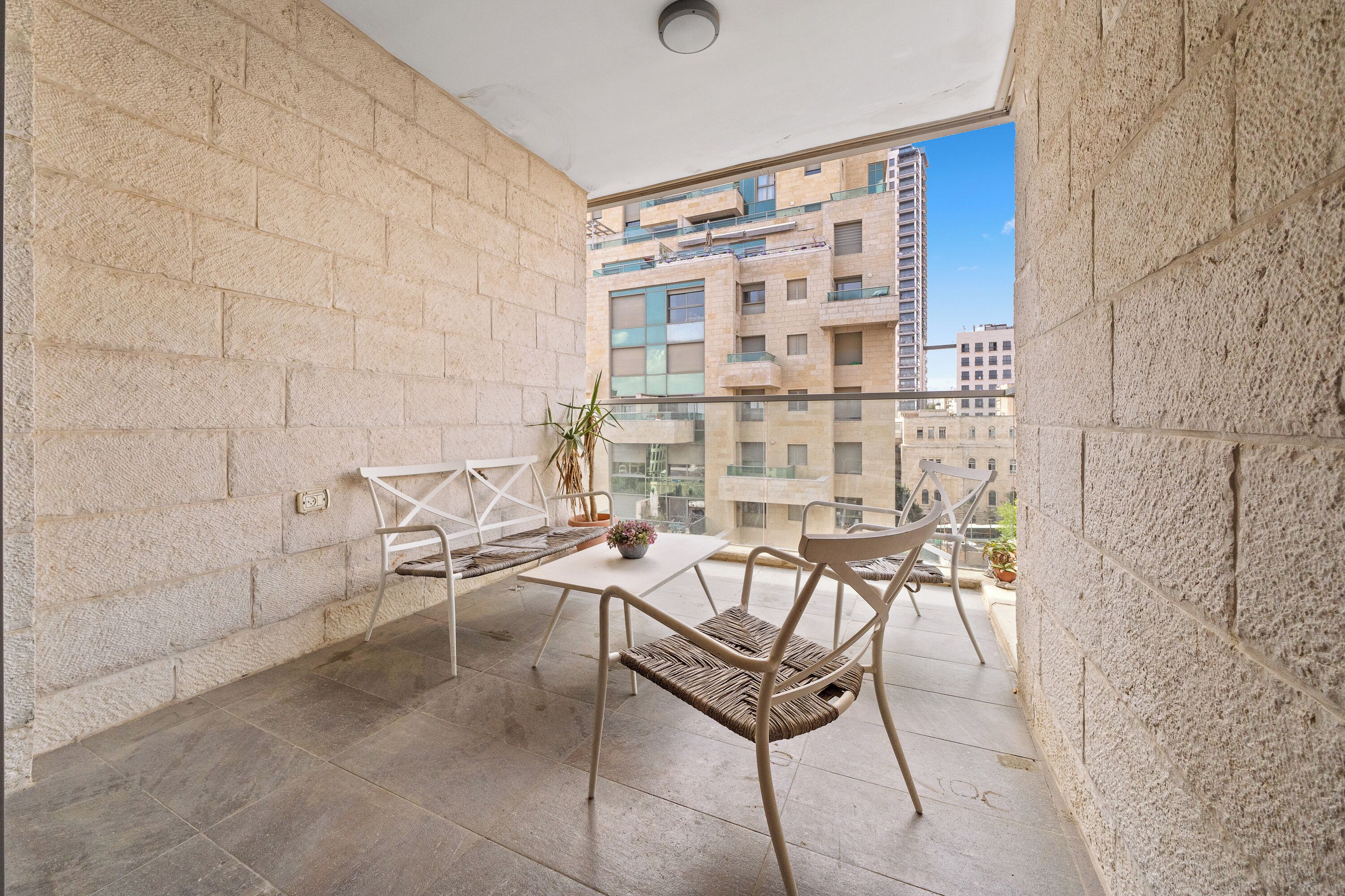 A balcony with a white table and wicker chairs, a potted plant, and a view of high-rise buildings.