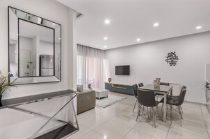 Una sala de estar moderna con comedor, un espejo grande, televisor de pantalla plana y cocina al fondo.