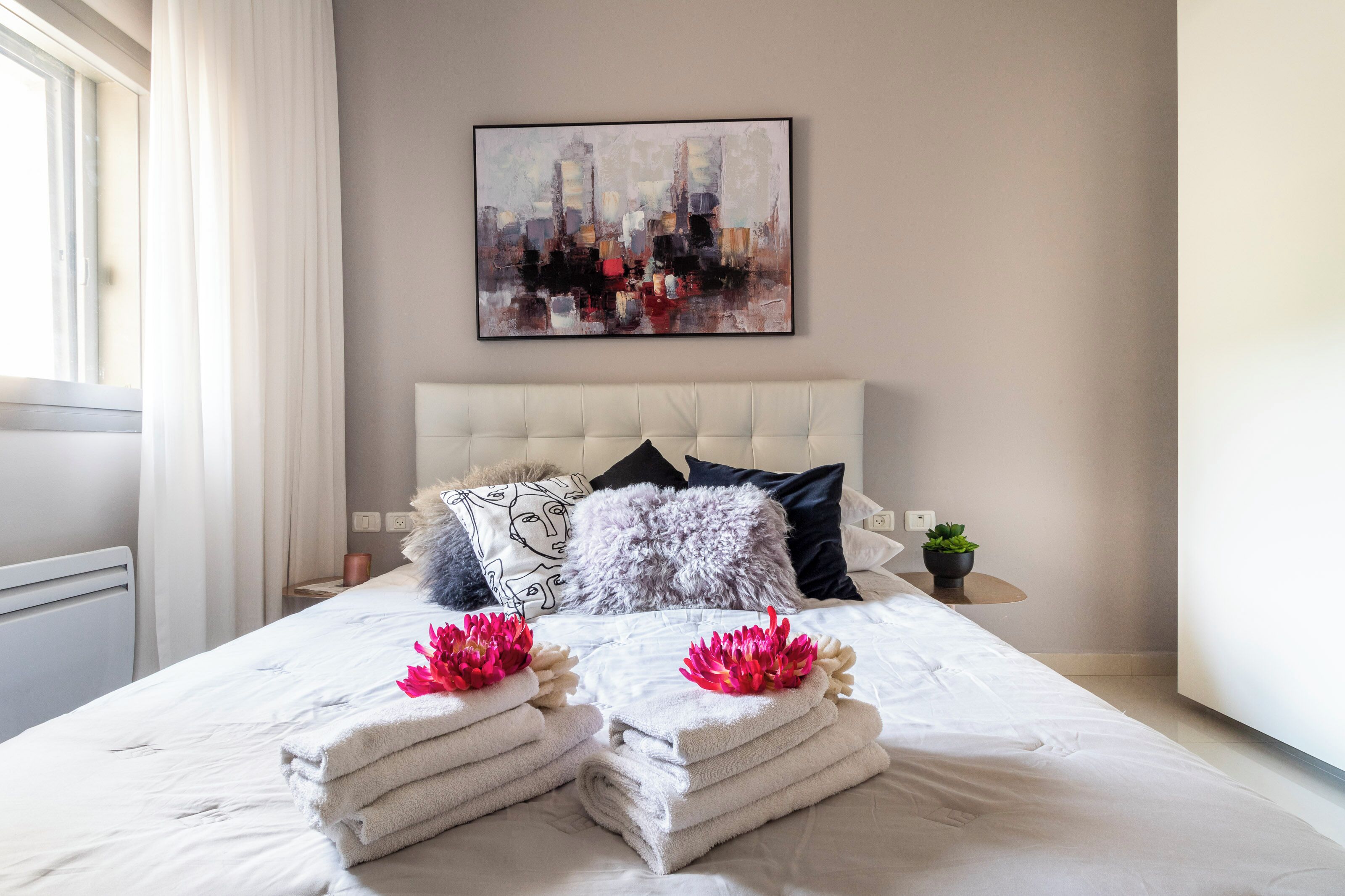 Un lit bien fait, avec du linge de lit blanc et des fleurs roses, un tableau représentant un paysage urbain accroché au mur, et une petite plante en pot posée sur la table de chevet.