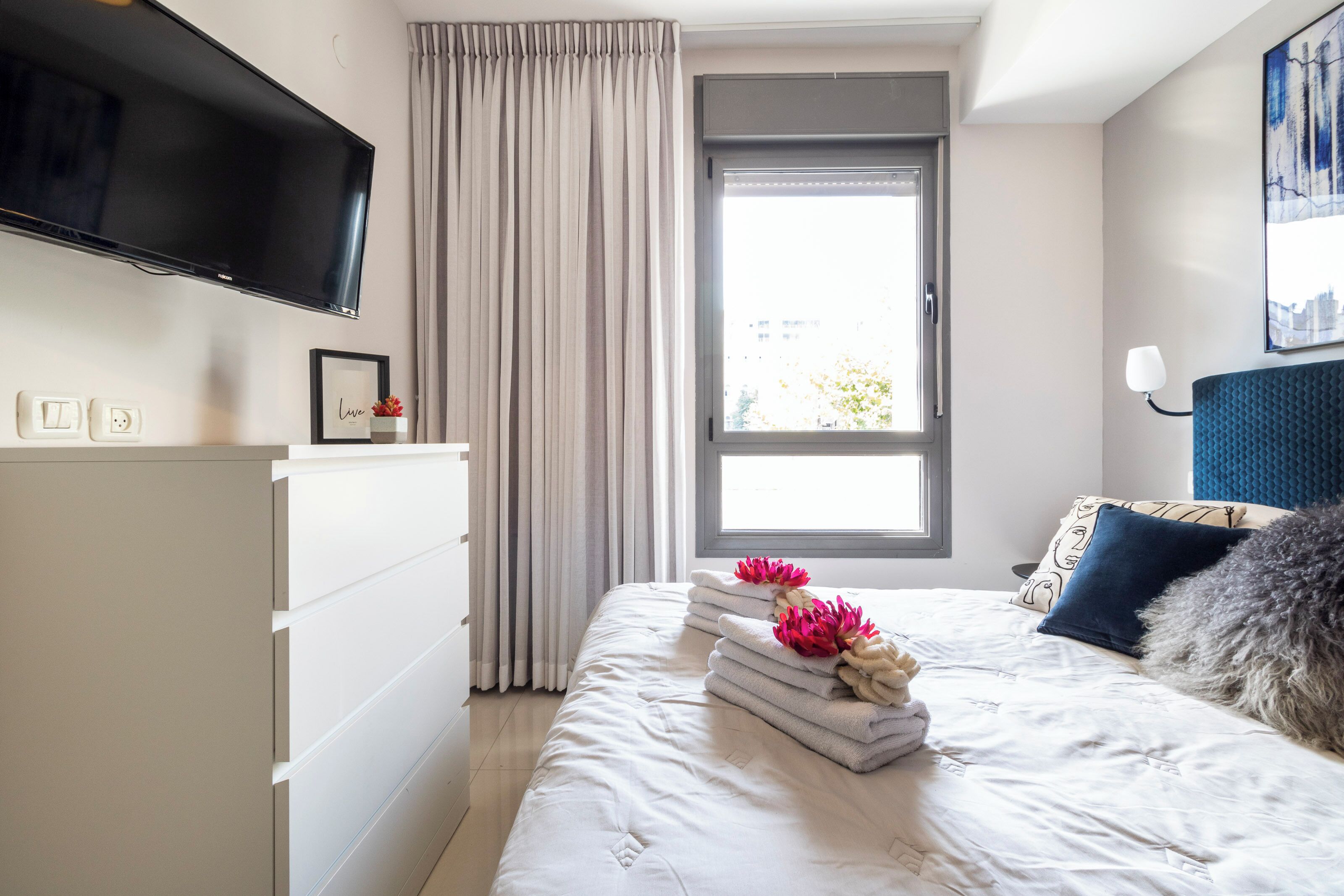 A hotel room with a bed, a TV mounted on the wall, a window with curtains, and a bedside table with a lamp.