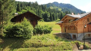 Una cabaña de madera con balcón, rodeada de exuberante vegetación y montañas al fondo.