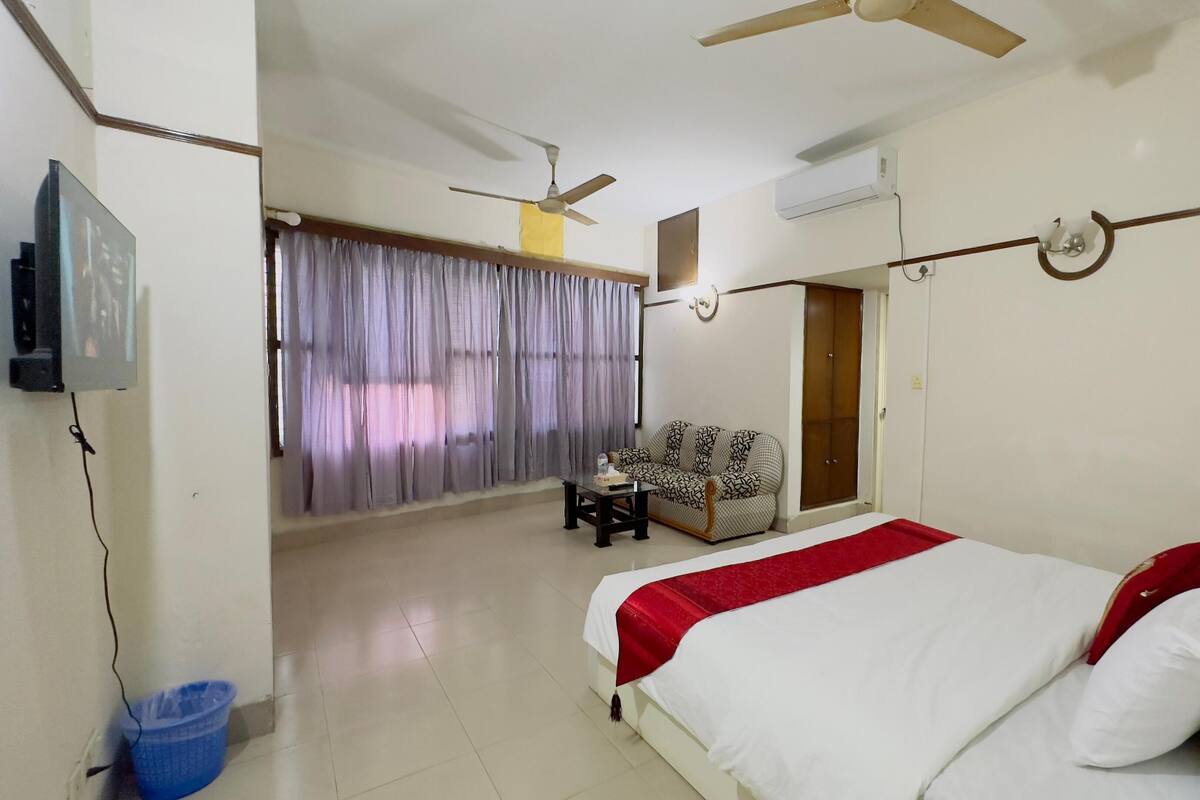 A hotel room with a bed, a sofa, a ceiling fan, an air conditioning unit, and a television.