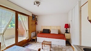 A compact bedroom with a bed, bedside table, and a small stool. A window with curtains is visible, and a door leads to an outdoor area.
