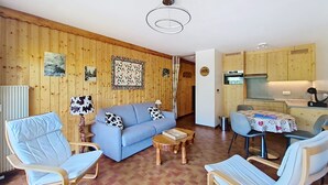A cozy room with a blue sofa, two armchairs, a small wooden table, and a kitchen area with wooden cabinets and a stove. - Champs Fleuris CFS7 COSY & VIEW 3 Pers. (Châtel)