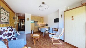 A compact living space with a sofa, a dining table, a chair, a kitchen area, and a TV mounted on the wall. - Champs Fleuris CFS7 COSY & VIEW 3 Pers. (Châtel)