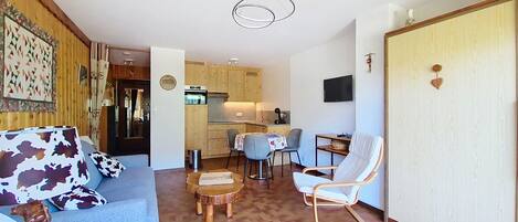 Ein kompakter Wohnraum mit Sofa, Esstisch, Stuhl, Küchenzeile und Wandfernseher.
