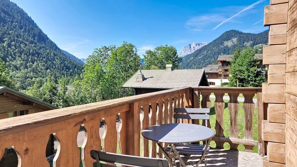 A wooden balcony with a table and chairs, overlooking a mountainous landscape with buildings in the distance.