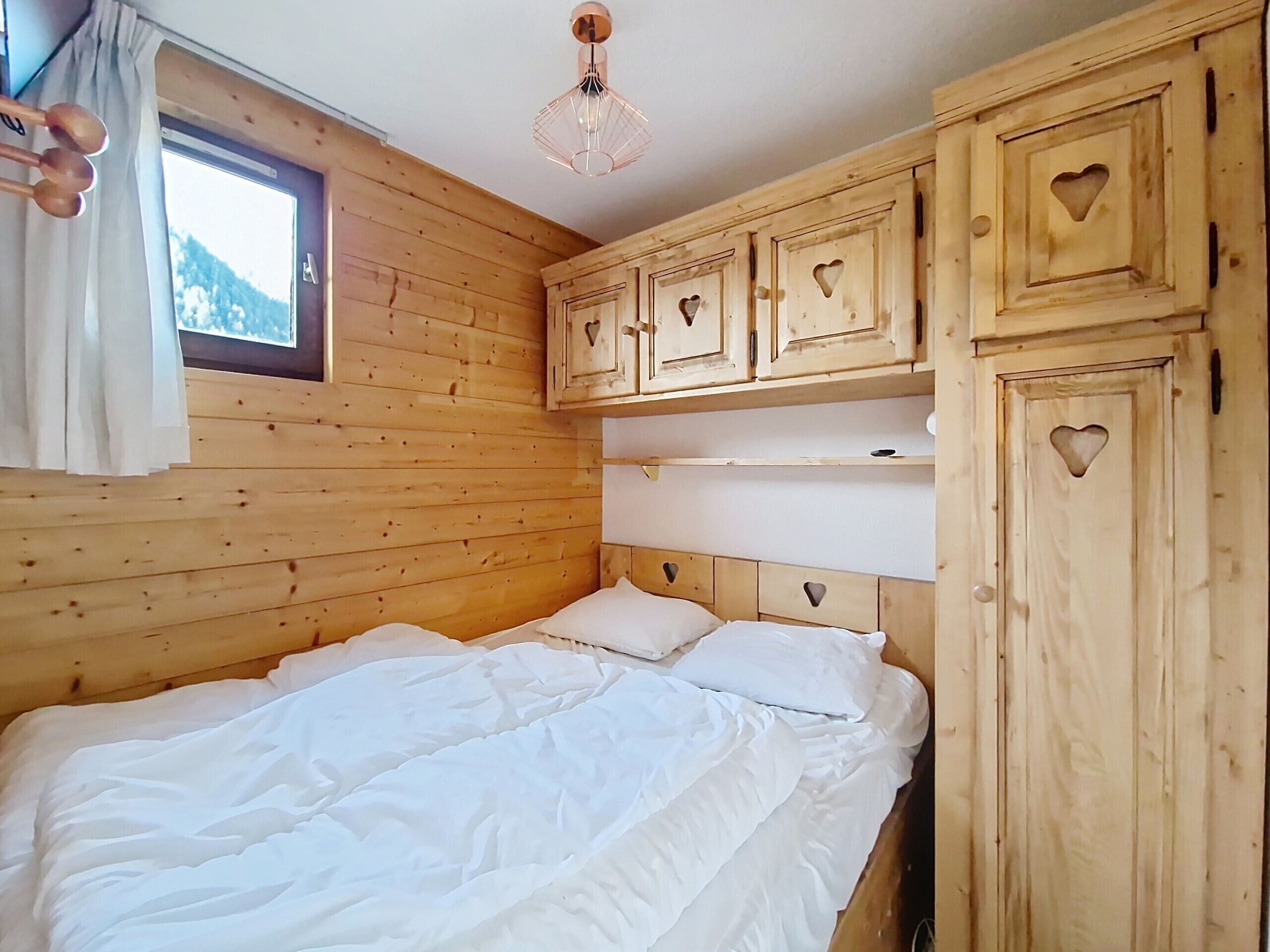 A wooden room with a bed, cabinets with heart-shaped cutouts, and a window with curtains.