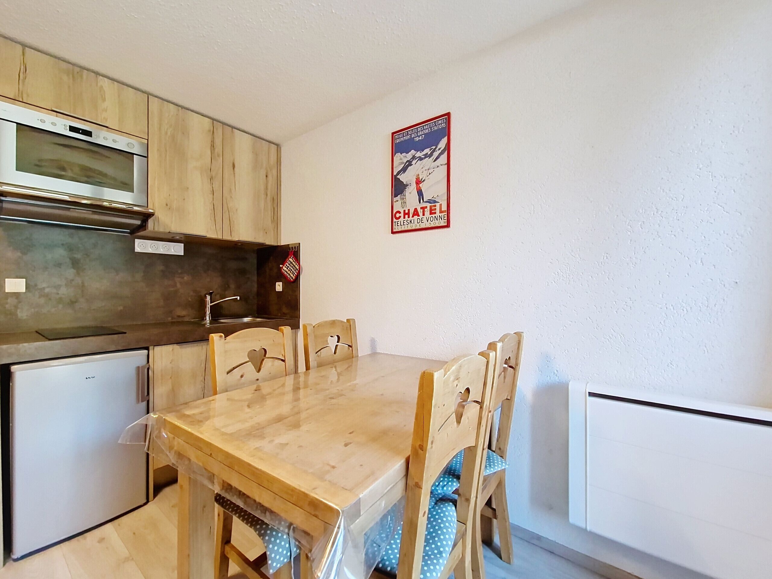 Une cuisine compacte avec des meubles en bois, un four encastré et un petit réfrigérateur. On y trouve une table à manger en bois avec quatre chaises, ainsi qu’une affiche accrochée au mur.