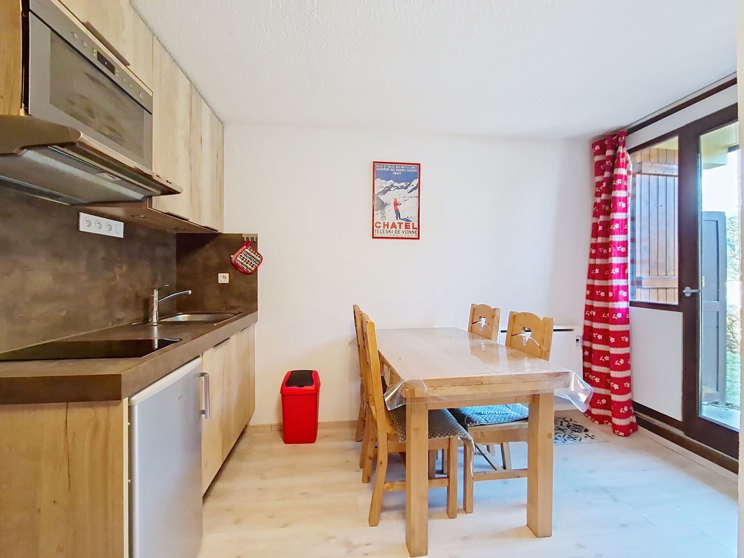 Eine kleine Küche mit Holzschränken, einer Spüle aus Edelstahl und einem Essbereich mit Tisch und Stühlen. Ein roter Mülleimer ist in der Nähe der Küche zu sehen, und ein Poster befindet sich an der Wand.
