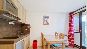Cocina compacta con armarios de madera, pileta de acero inoxidable y comedor con mesa y sillas. Se observa un cubo de basura rojo cerca de la cocina, y un cartel en la pared.
