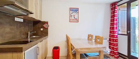 Eine kleine Küche mit Holzschränken, einer Spüle aus Edelstahl und einem Essbereich mit Tisch und Stühlen. Ein roter Mülleimer ist in der Nähe der Küche zu sehen, und ein Poster befindet sich an der Wand.
