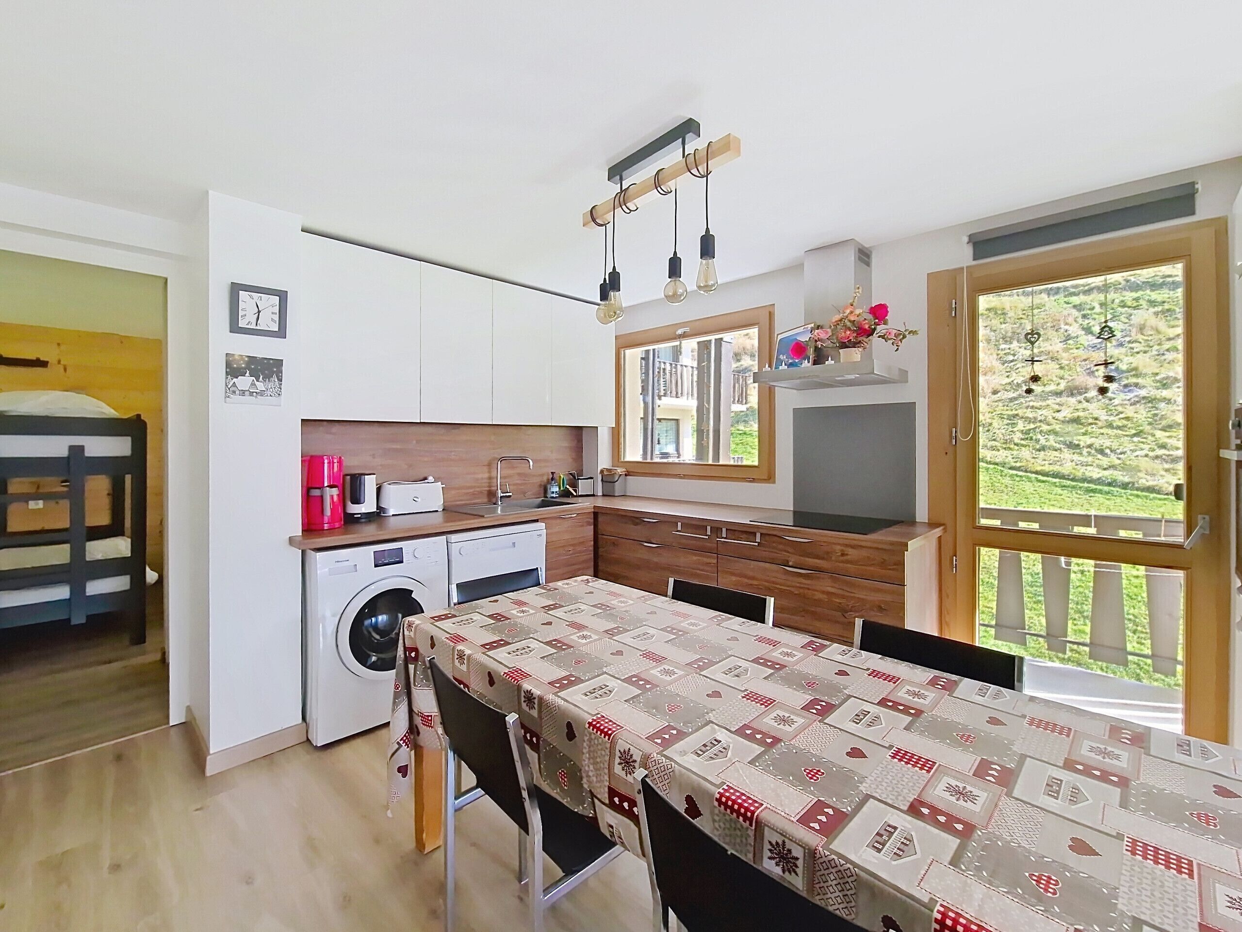 A compact kitchen with a washing machine, a dining table with a patterned tablecloth, and a window with a view of greenery.