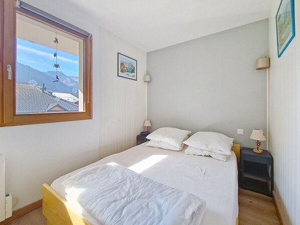 A bedroom with a bed, two bedside tables, a window with a view of buildings and mountains, and a framed picture on the wall.