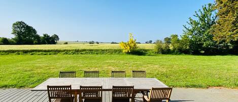 Een houten terras met een tafel en stoelen, uitkijkend over een grasveld met bomen en een heldere lucht.
