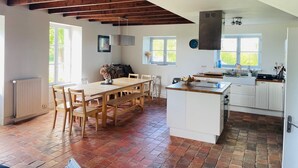 A spacious kitchen with a large island, wooden beams on the ceiling, and a view of the outdoors through the windows. - Grande Maison de Vacances et WE Près de Dinan (Trévron)