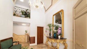 Een statige hal met een marmeren vloer, een groene bekleden stoel, een gouden console tafel met een spiegel, en een trap met een balustrade.