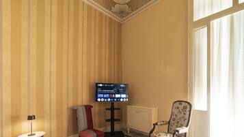 Ein Hotelzimmer mit einem an der Wand befestigten Fernseher, einem Sessel mit floralem Bezug und einem Fenster mit transparenten Vorhängen.