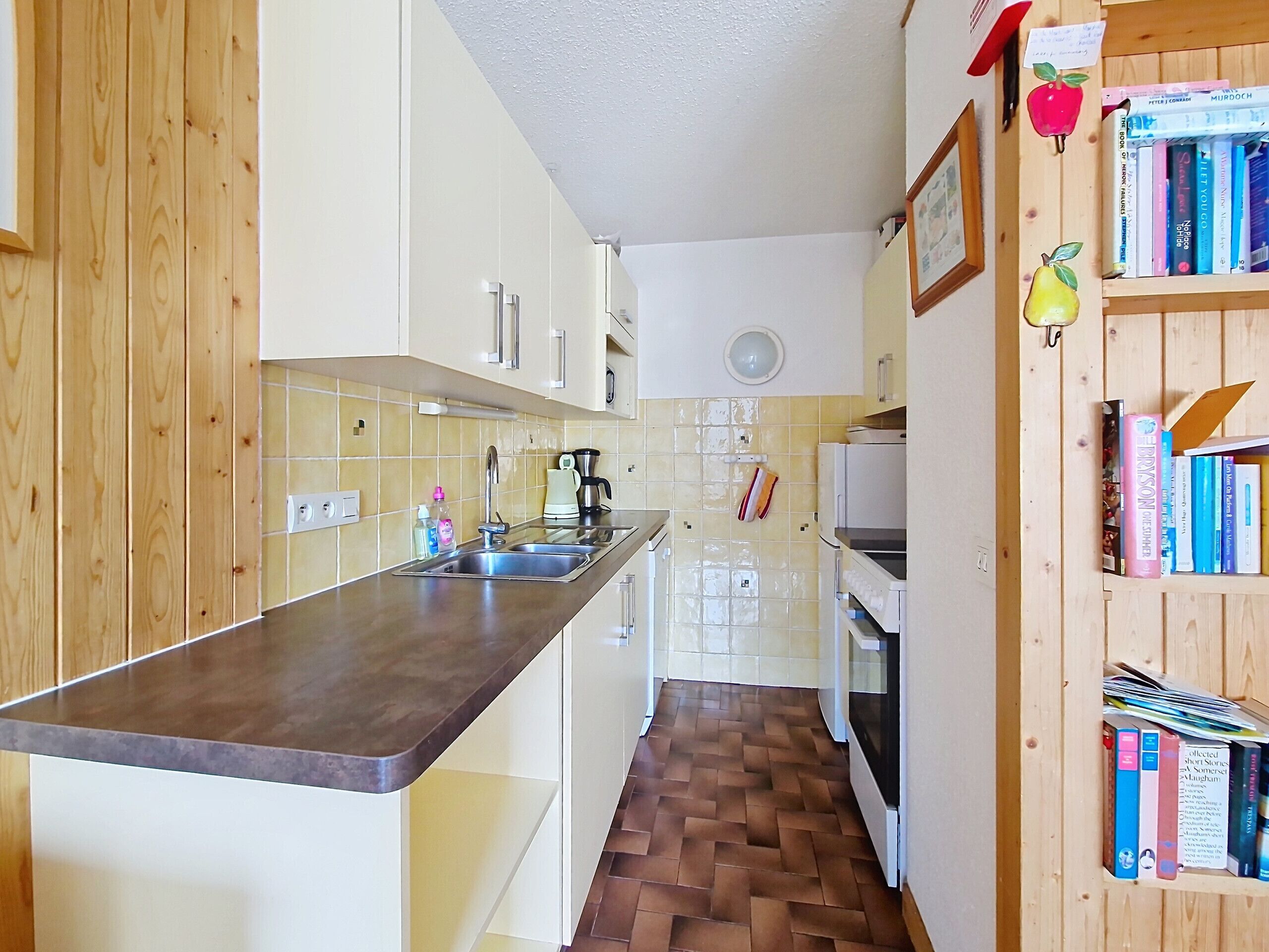 A kitchen with wooden cabinets, a countertop, a sink, a stove, and a bookshelf with books.