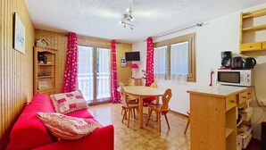 A compact living space with a red sofa, a dining table with chairs, a kitchenette with a microwave, and a television on the wall. - Gelinote GL3 TELECABINE & MOUNTAIN 4 Pers. (Châtel)