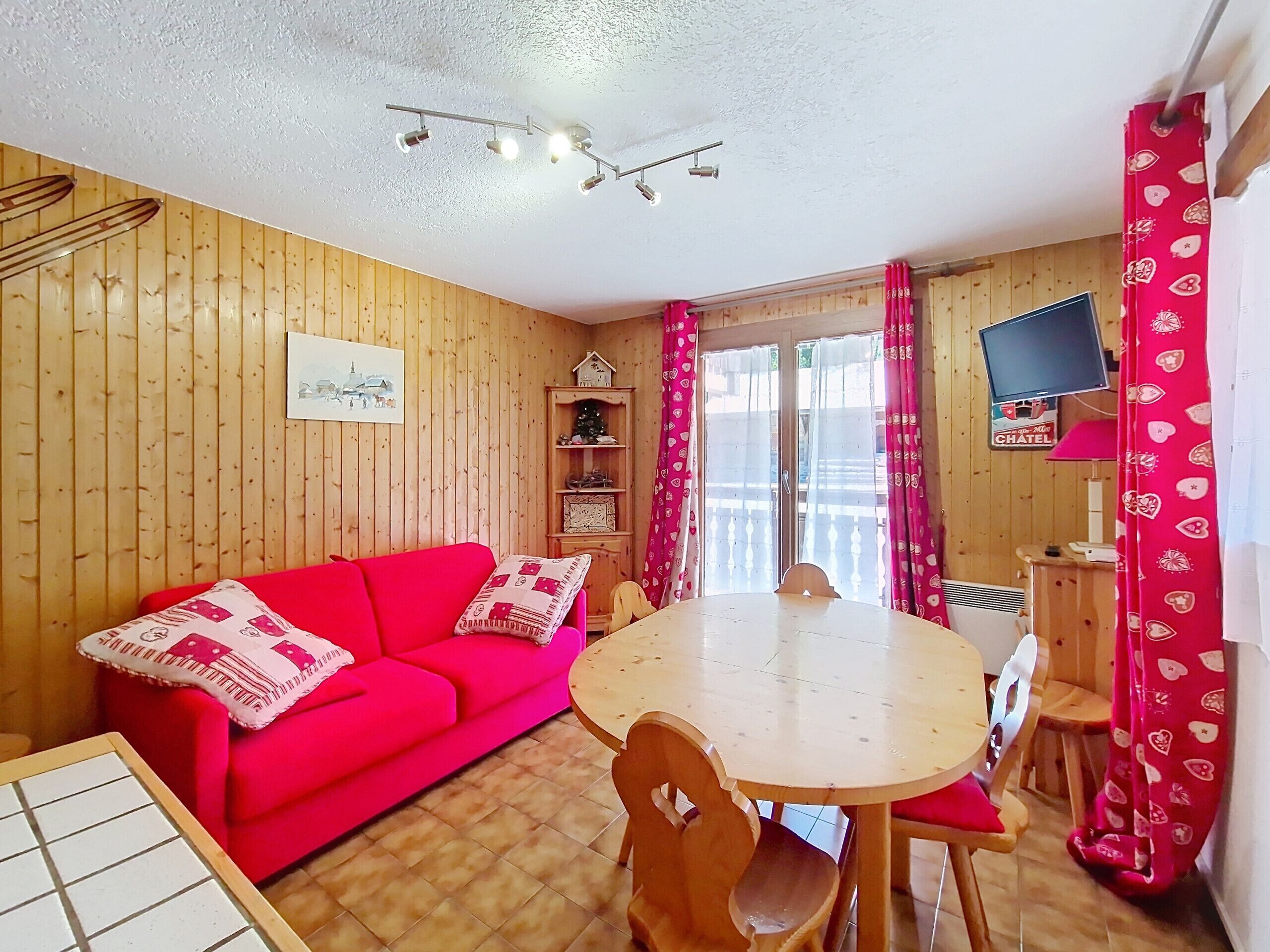 Ein gemütliches Zimmer mit einem roten Sofa, Holzwänden, einem runden Tisch mit Stühlen, einem Fernseher an der Wand und einem Fenster mit Vorhängen.