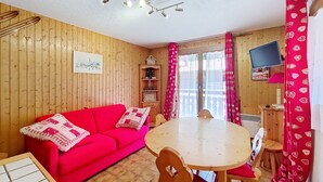 A cozy room with a red sofa, wooden walls, a round table with chairs, a TV on the wall, and a window with curtains. - Gelinote GL3 TELECABINE & MOUNTAIN 4 Pers. (Châtel)