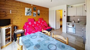 A cozy room with a wooden wall, a bed with red bedding, a dining table with a blue tablecloth, and a kitchen area with modern appliances. - Corina CN11 QUIET & MOUNTAIN 4 Pers. (Châtel)