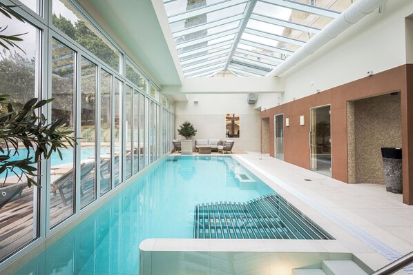 An indoor swimming pool with clear glass walls, a seating area with a sofa and chairs, and a cross on the wall.