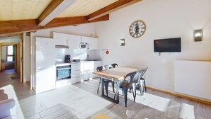 A modern kitchen with white cabinets, a microwave, and a coffee maker. There is a dining area with a wooden table and chairs. A clock is on the wall, and a flat-screen TV is mounted above the table.