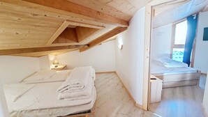 A room with a loft bed, wooden ceiling beams, a small bedside table with a lamp, and a view of another room through an open door.